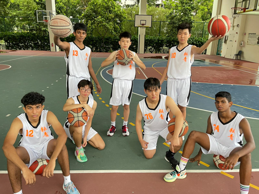 Photograph of a basketball team posing on an outdoor court, featuring seven players wearing white jerseys with orange numbers and logos. Players hold basketballs in various poses, with numbers ranging from 10 to 76 visible, and the background includes basketball hoops and greenery.
